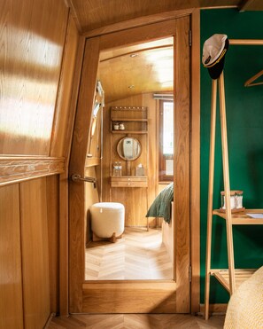 Un interior de madera con una puerta que da al baño, un espejo redondo y una estantería con artículos de aseo.