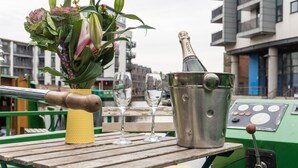 A table with a champagne bottle, glasses, and a vase of flowers on a boat. - 4* Luxury floating wide beam houseboat moored in Edinburgh city center. (Edinburgh)