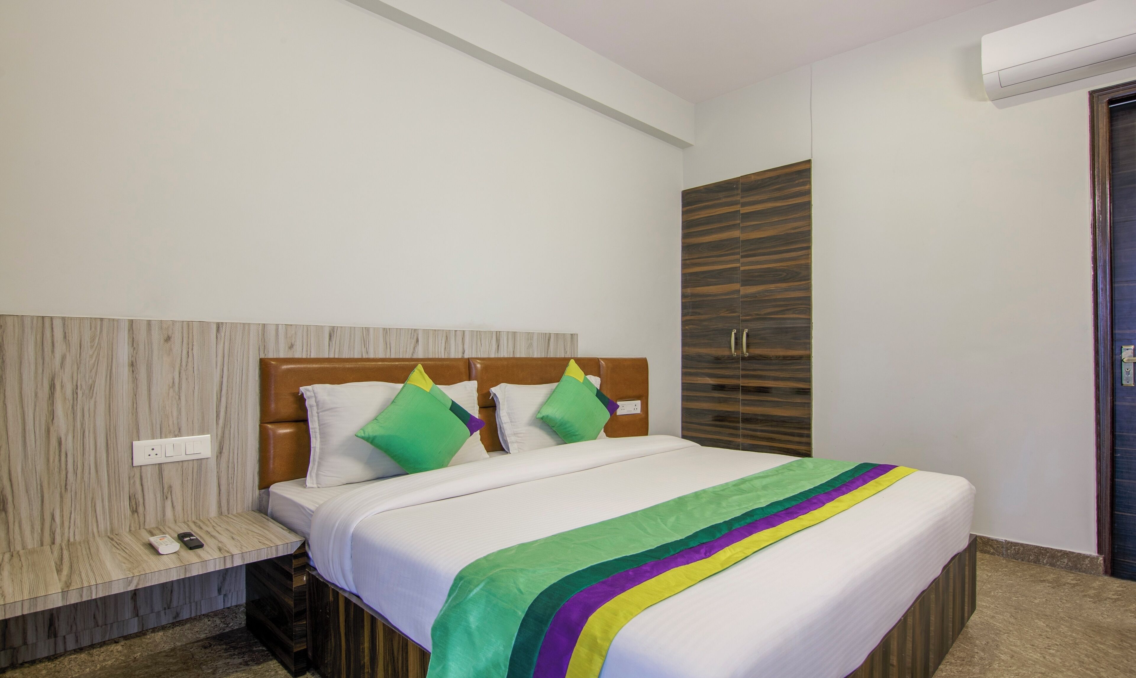 A hotel room with a bed, a wooden wardrobe, a bench with a phone and remote, and an air conditioning unit.