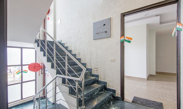 Un escalier avec des marches en marbre et des rampes en métal, menant à une pièce dont le mur est blanc et qui possède une porte ornée de drapeaux indiens.