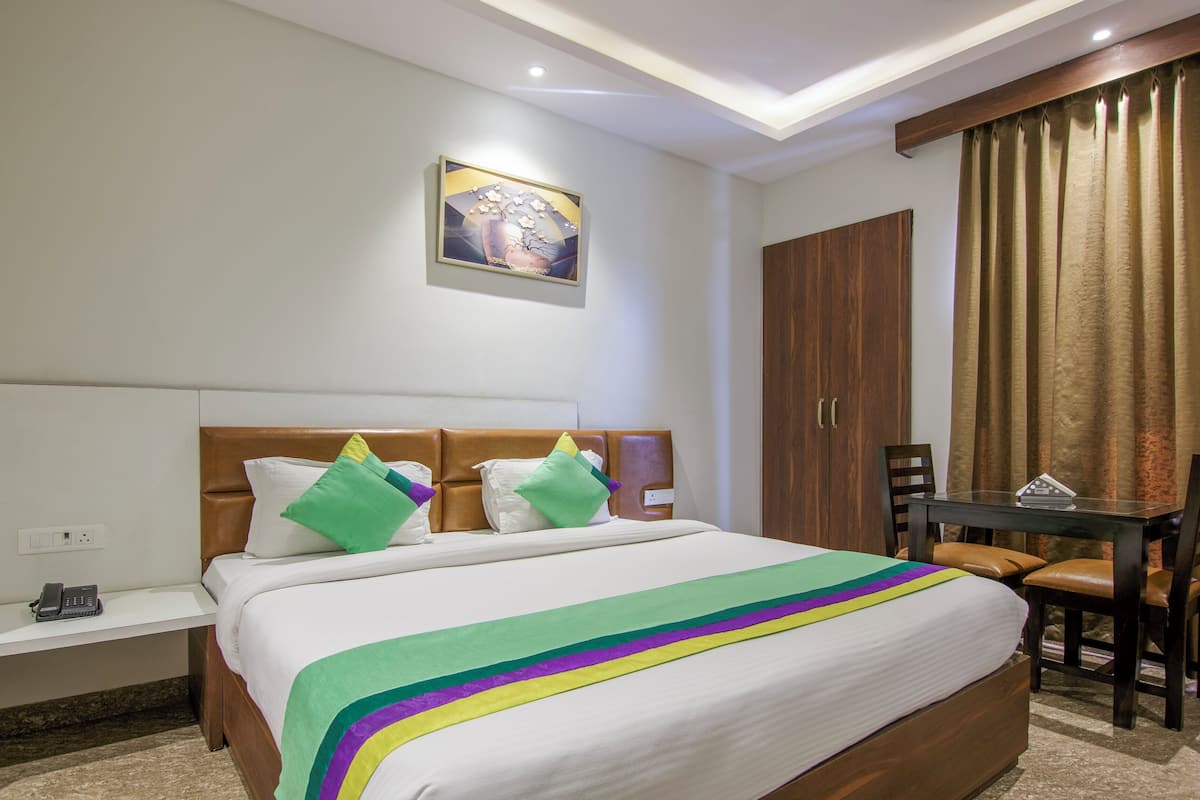 A hotel room with a bed, bedside table, a desk with a chair, a wardrobe, and a painting on the wall.