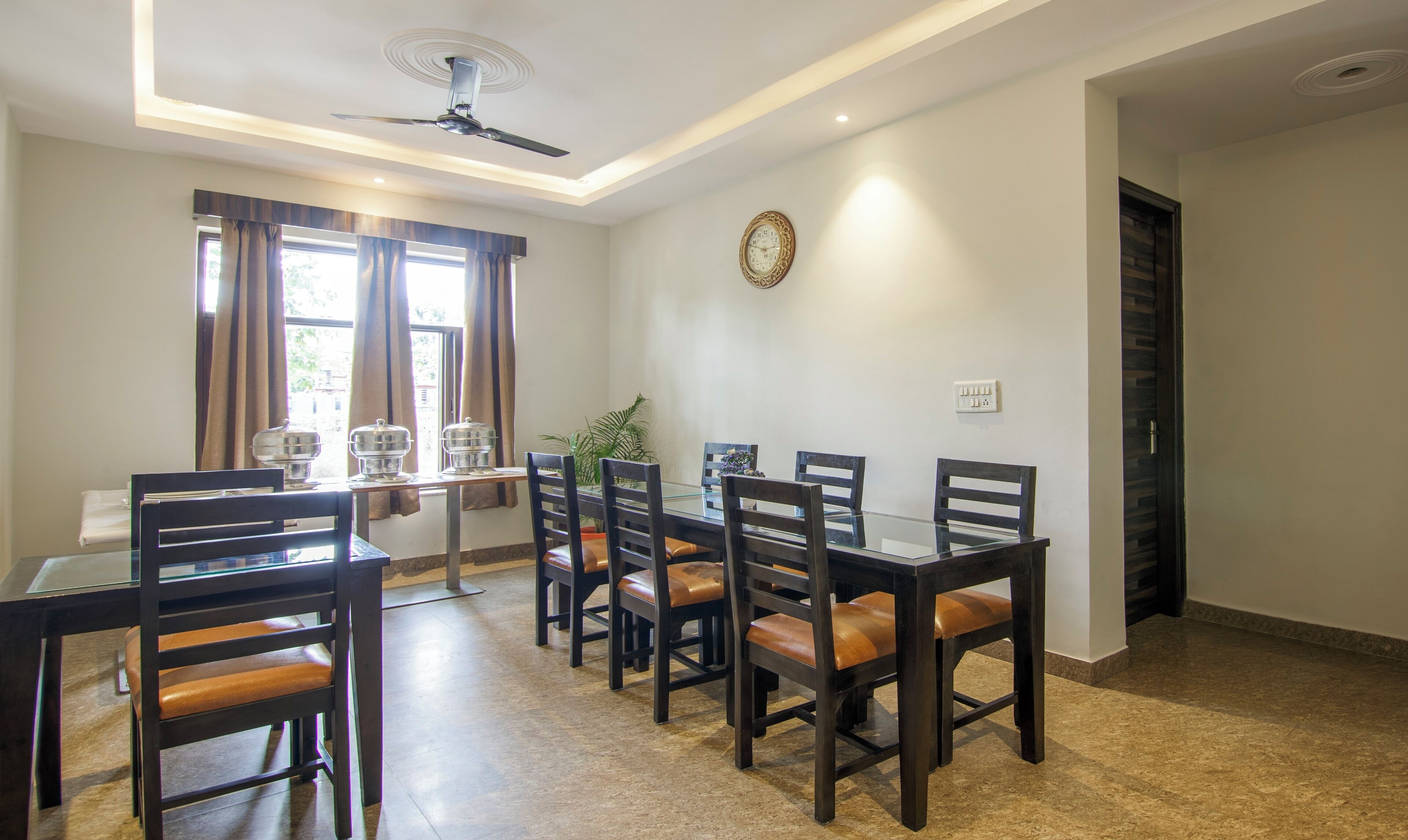 Un comedor con una mesa de vidrio, sillas de madera, un ventilador de techo y un reloj de pared.