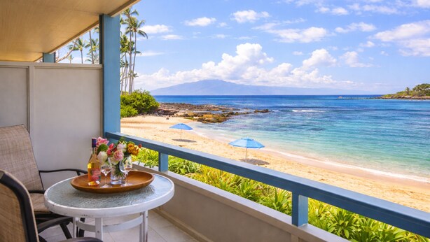 Un balcon avec une table, des chaises et une vue sur une plage à l’eau claire, avec une montagne qui se profile à l’horizon.