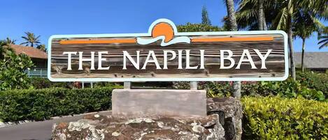 Een houten bord met 'THE NAPI LI BAY' erop, gemonteerd op een stenen basis, met een palmboom en begroeiing op de achtergrond.