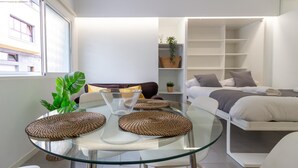 Ett modernt och kompakt vardagsrum med ett glasbord, en soffa, en säng och en liten hylla.