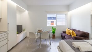 Ett kompakt hotellrum med en säng, en soffa, ett matbord med stolar, en TV och ett fönster med utsikt över en byggnad.