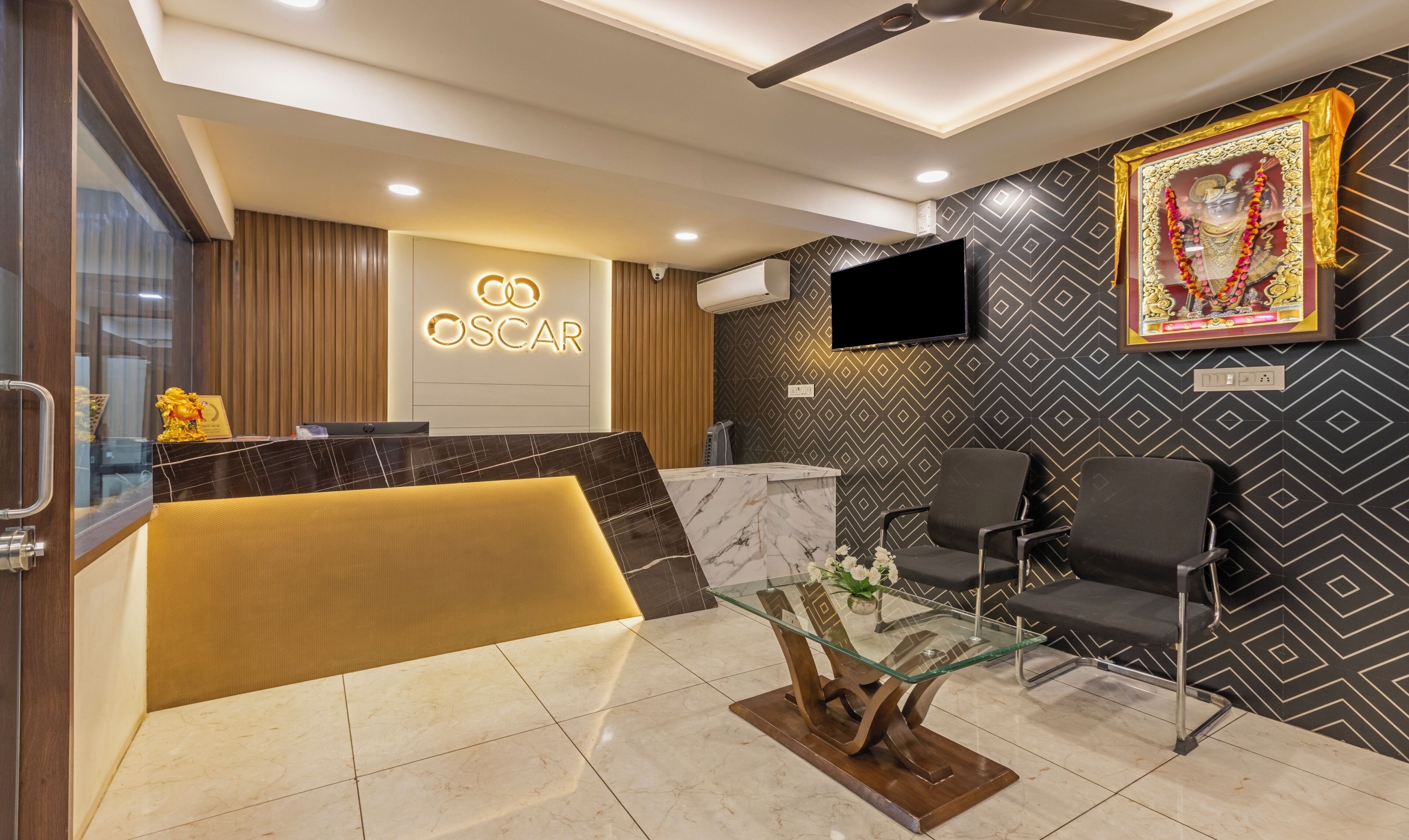A hotel reception area with a marble reception desk, a sofa, and a glass coffee table. There is a television mounted on the wall and a framed picture above the reception desk.