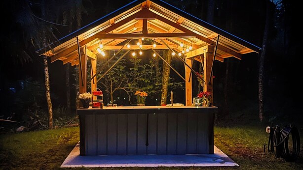 Un pabellón de madera con una plataforma elevada, luces colgantes y plantas en macetas.