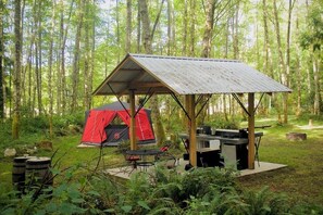 Un terrain de camping avec une tente rouge, une table de pique-nique couverte et un espace barbecue.