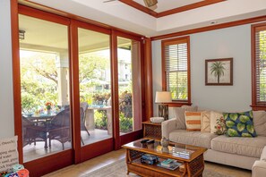 A living room with a sofa, coffee table, and a view of the outdoors through large sliding glass doors. - Kolea - Walk to Beach with Pool Access (Waikoloa)