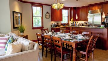 A dining area with a wooden table set for four, surrounded by chairs. A kitchen with wooden cabinets and a stainless steel refrigerator is visible in the background.