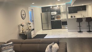 A modern living room with a brown sectional sofa, a small white dog, and a kitchen in the background with dark cabinets and stainless steel appliances. - Modern Spacious 1 bedroom and a den home (Lower Unit) (Windsor)