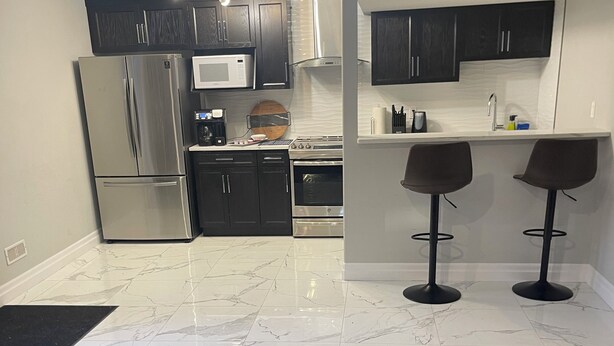 A modern kitchen with dark wood cabinets, stainless steel appliances, and a marble backsplash. There are two bar stools and a small island with a sink. - Modern Spacious 1 bedroom and a den home (Lower Unit) (Windsor)