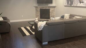 A modern living room with a sectional sofa, a flat-screen TV above a fireplace, and a large abstract painting on the wall. - Modern Spacious 1 bedroom and a den home (Lower Unit) (Windsor)