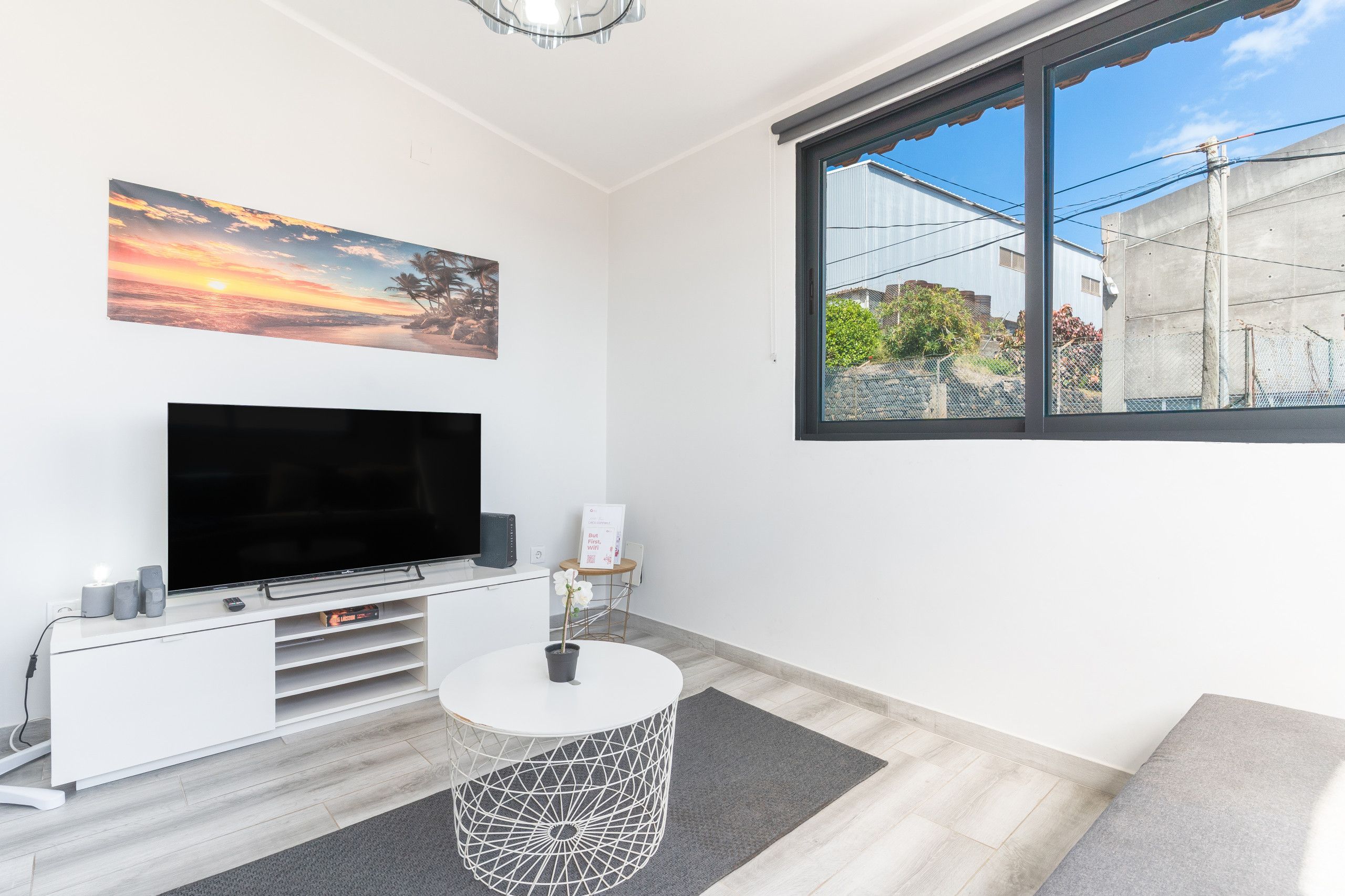 A modern living room with a flat-screen TV, a white console table, a round coffee table, and a large window with a view of buildings and greenery.