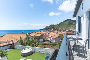 A balcony with a view of a coastal town, a hot tub, and a table with chairs.