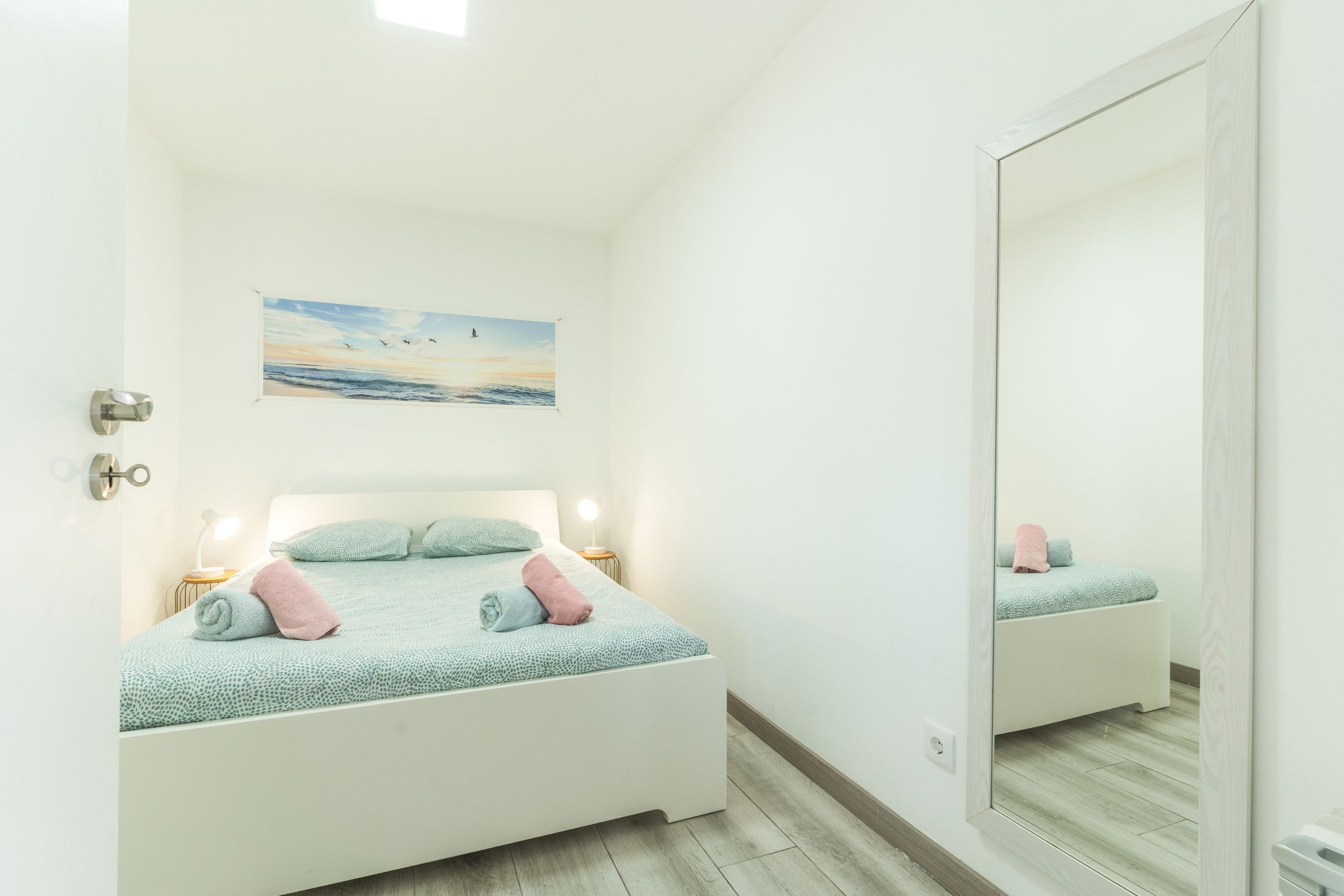 A bedroom with a bed, pillows, a nightstand, a lamp, a painting, and a mirror.