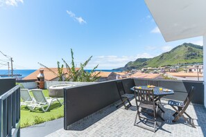 A balcony with a table and chairs, overlooking a coastal town and the sea.