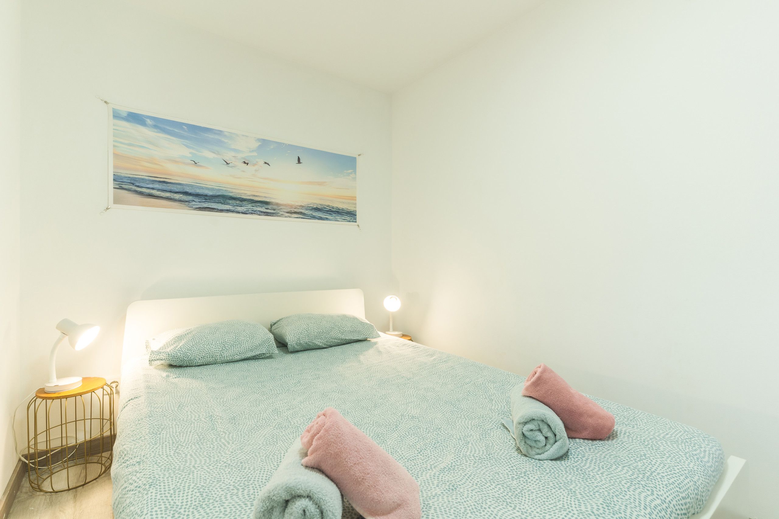 Quarto com cama, travesseiros, mesa de cabeceira, luminária e uma pintura de uma praia.
