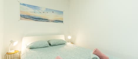 Une chambre à coucher avec un lit, des oreillers, une table de chevet, une lampe et un tableau représentant une scène de plage.