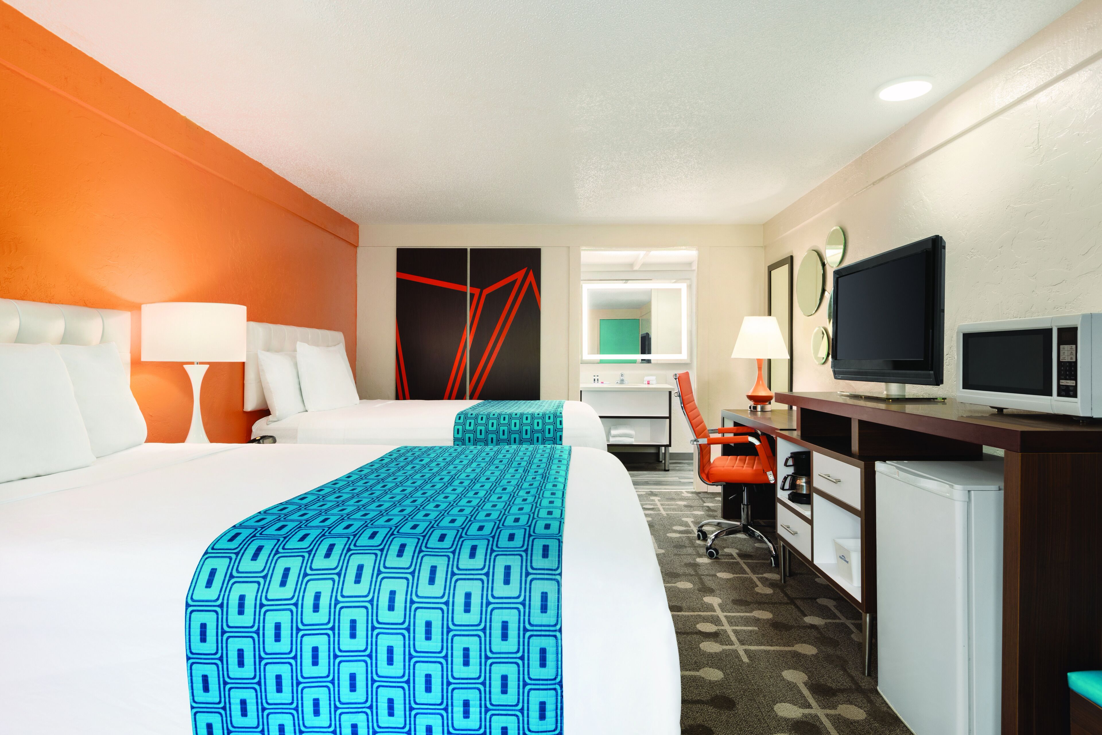 A hotel room with a large bed, a desk with a TV, a small fridge, and a bathroom visible through a window.