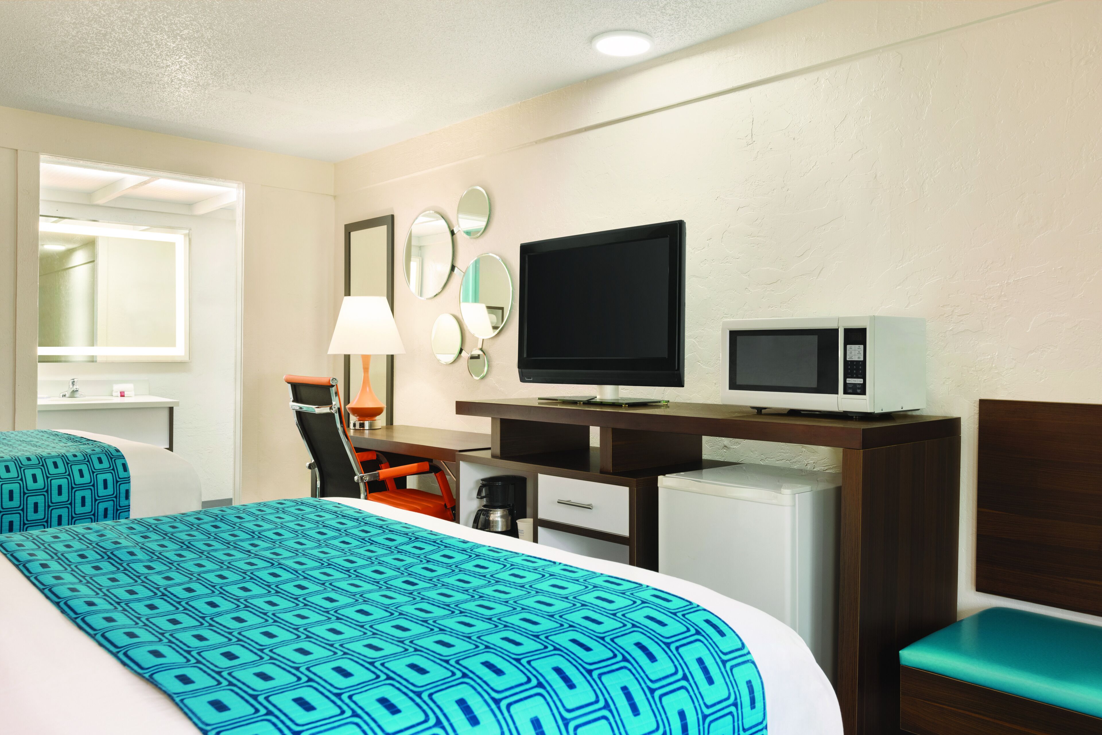 A hotel room with a bed, a desk with a TV and microwave, a chair, and a small refrigerator.