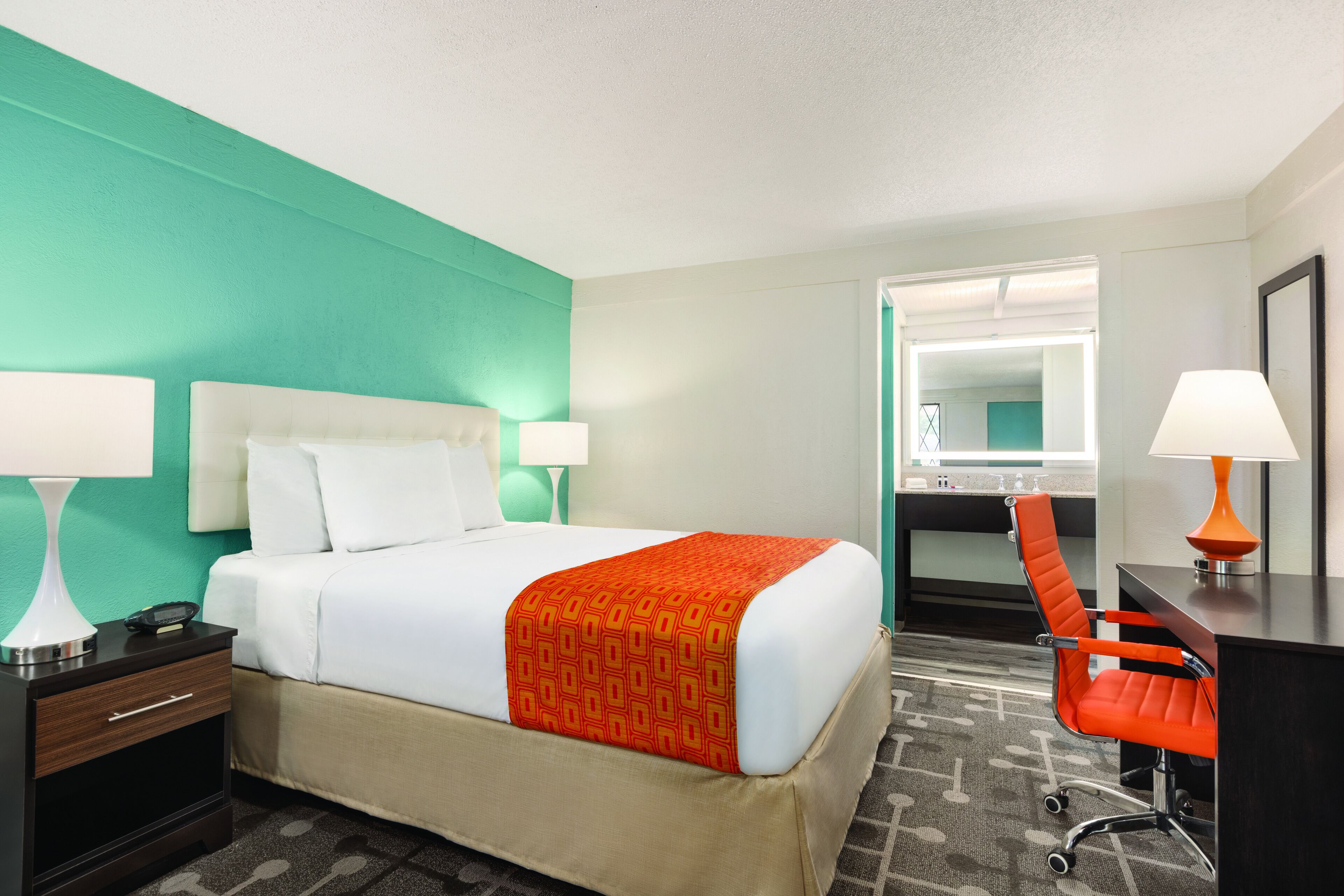 A hotel room with a bed, nightstand, desk, chair, and a bathroom visible through an open door.