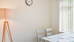 Ein Zimmer mit einem Tisch, der mit einer punktierten Tischdecke bedeckt ist, zwei weiße Stühle, einer Stehlampe aus Holz und einer Wanduhr.