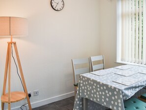 Une pièce avec une table recouverte d’une nappe à pois, deux chaises blanches, un lampadaire en bois et une horloge murale.