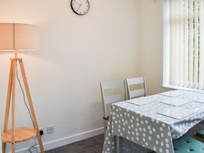 Una habitación con una mesa cubierta por un mantel a lunares, dos sillas blancas, una lámpara de pie de madera y un reloj de pared.