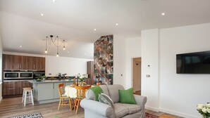 Una sala de estar moderna con un sofá gris, un comedor con mesa y sillas, una cocina con electrodomésticos integrados y un televisor montado en la pared.