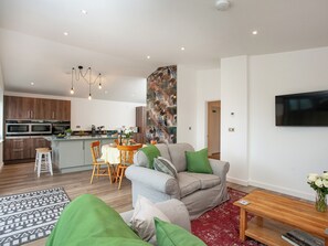 Una sala de estar moderna con un sofá gris, un comedor con mesa y sillas, una cocina con electrodomésticos integrados y un televisor montado en la pared.