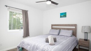 Ein Schlafzimmer mit einem Bett, einem Nachttisch, einer Lampe, einem Deckenventilator, einem Fenster mit Vorhängen und einem Bild an der Wand.