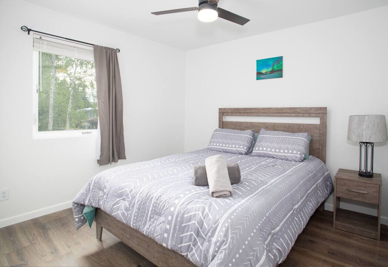 Une chambre à coucher avec un lit, une table de chevet, un ventilateur de plafond, une fenêtre avec des rideaux et un tableau accroché au mur.
