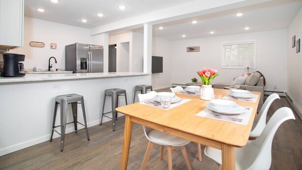 A modern kitchen with white cabinets, a stainless steel refrigerator, and a dining area with a wooden table and white chairs. - 3b2b newly remodeled modern Fairbanks house close to everything (Fairbanks)
