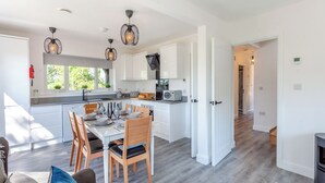 Eine moderne Küche mit weißen Schränken, ein Essbereich mit Tisch für vier Personen und ein Wohnzimmer mit Sofa und blauem Teppich.