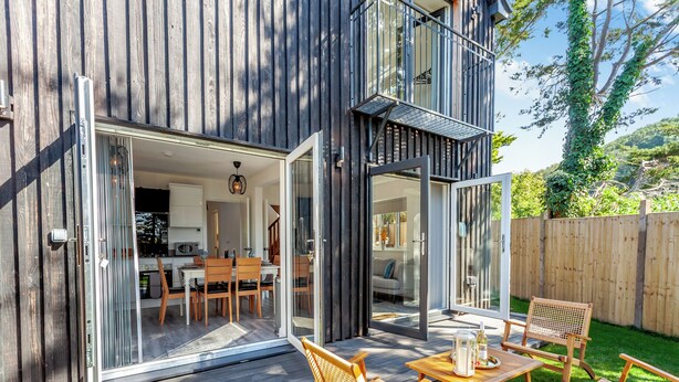 Ein modernes Haus mit einer Terrasse, Sitzgelegenheiten im Freien und einer Glastür, die zu einem Essbereich führt.