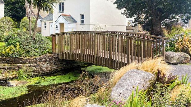 Una casa blanca con techo inclinado, un puente de madera sobre un arroyo pequeño y abundante vegetación alrededor.