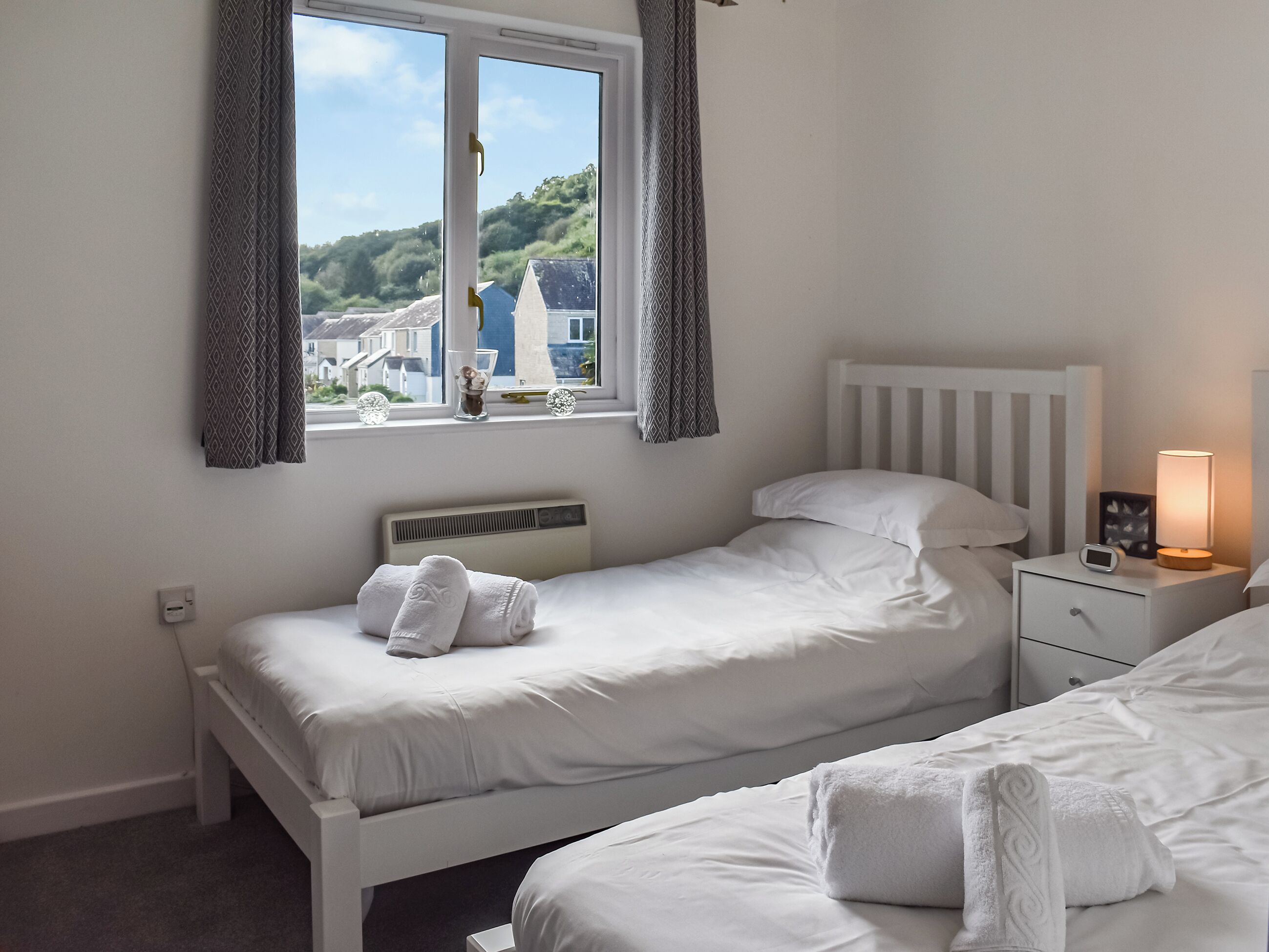 A bedroom with two beds, a window with curtains, a bedside table with a lamp, and a thermostat.