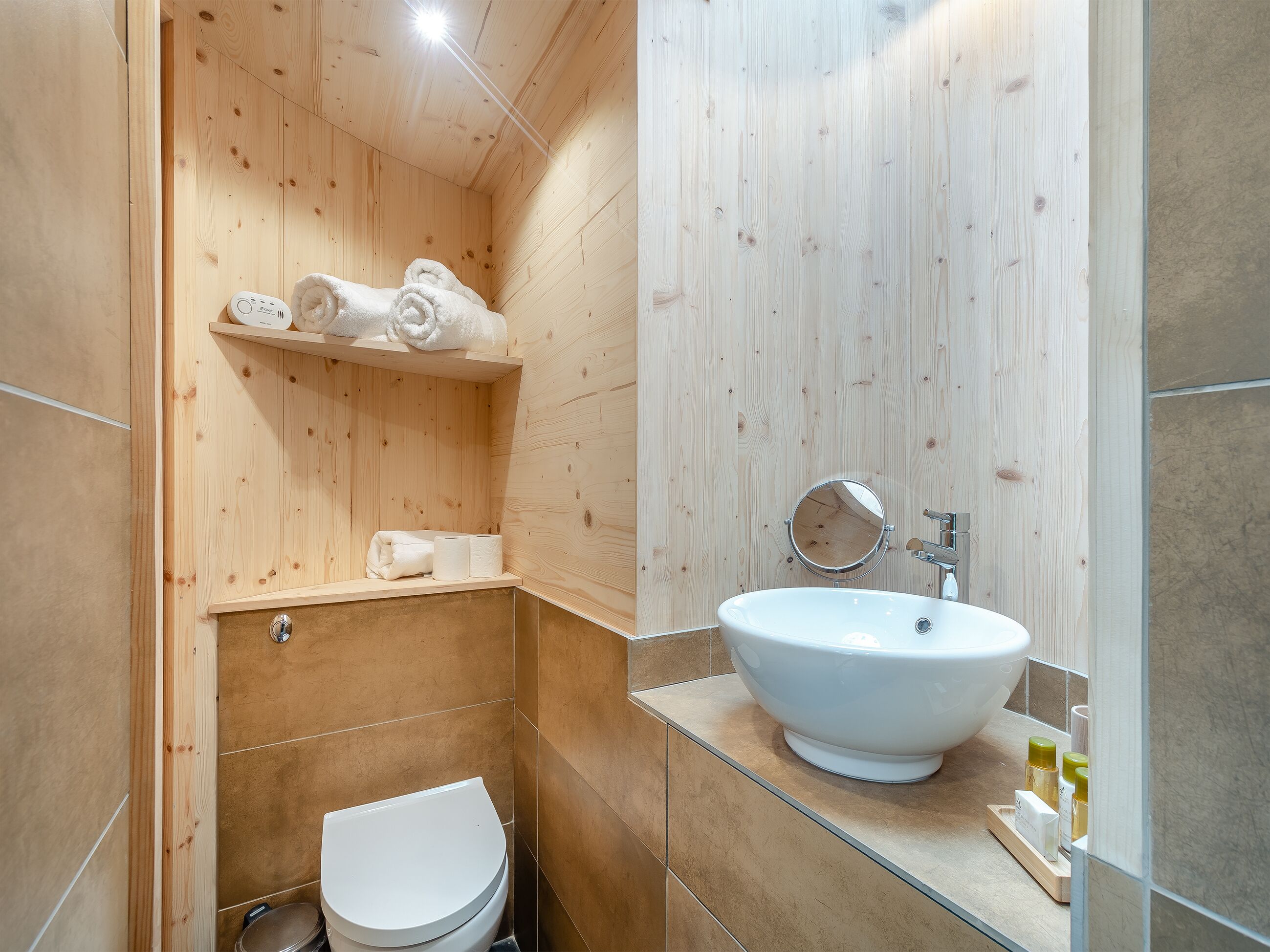 Un baño con inodoro blanco, estante de madera con toallas, espejo redondo y lavamanos blanco con encimera de vidrio.