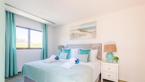 A bedroom with a large bed, teal curtains, a painting on the wall, and a nightstand with a lamp and a vase. - Falesia Beach C at Falesia Beach (Albufeira)