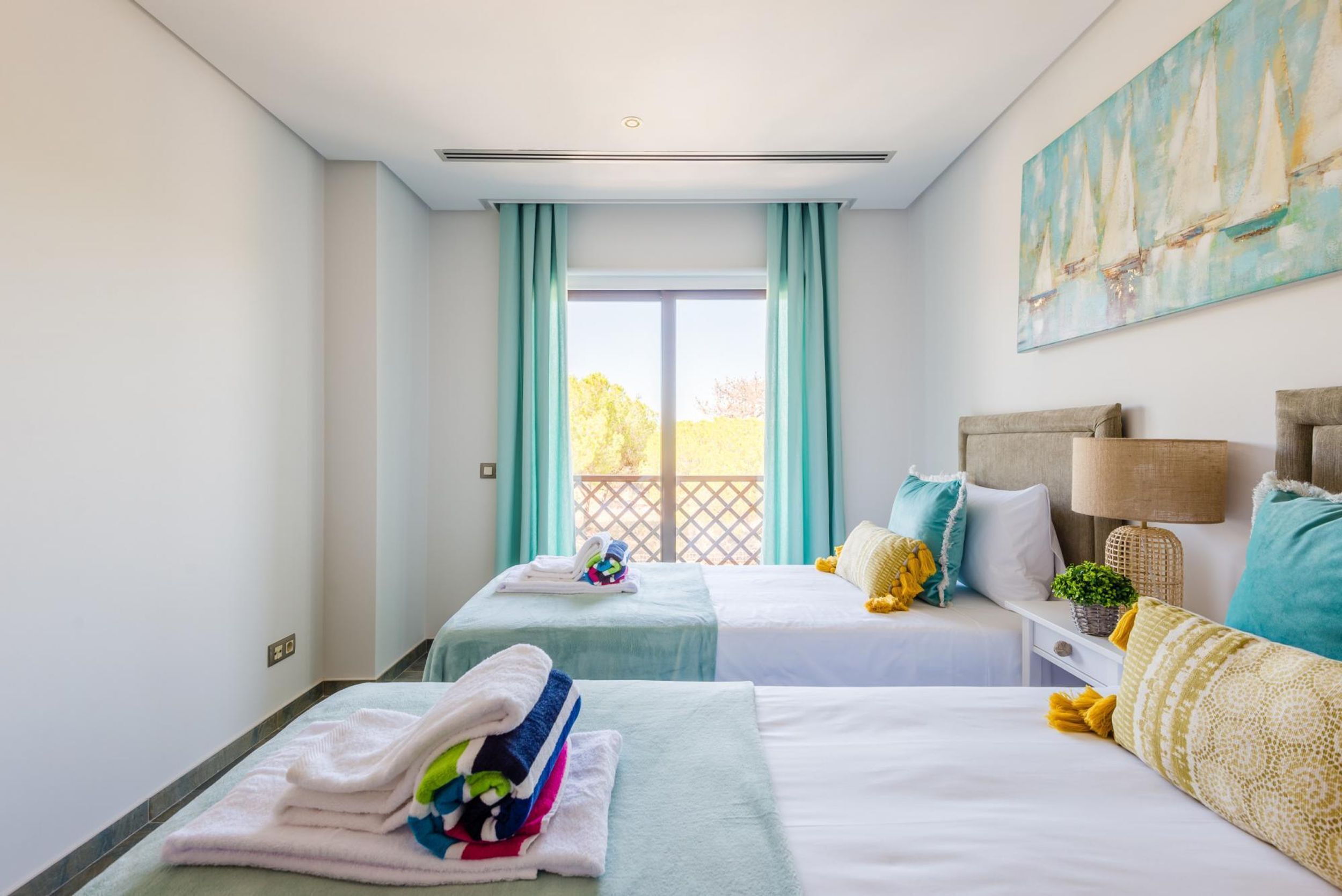 Une chambre d’hôtel avec deux lits, une grande fenêtre avec des rideaux, un tableau au mur et une petite table avec une lampe.