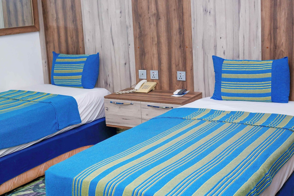 Une chambre d’hôtel avec deux lits, chacun recouvert d’une literie à rayures bleues et vertes. On y trouve une tête de lit en bois, une table de chevet en bois avec un téléphone et une calculatrice, ainsi qu’une commode en bois avec des tiroirs.