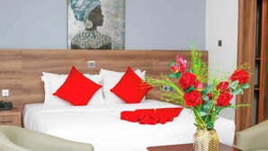 A hotel room with a wooden headboard, white bedding, red pillows, a wooden nightstand with a phone, a beige armchair, a wooden side table with a vase of red roses, and a painting on the wall. - Ave Maria Hotel (Accra)