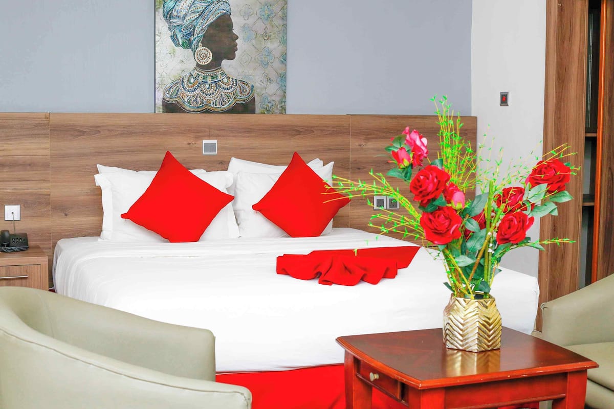 Une chambre d’hôtel avec une tête de lit en bois, une literie blanche, des coussins rouges, une table de chevet en bois avec un téléphone, un fauteuil beige, une table d’appoint en bois avec un vase de roses rouges et un tableau au mur.