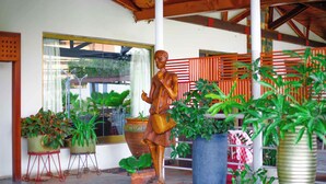 A wooden statue in a covered outdoor area with potted plants and a large window. - Ave Maria Hotel (Accra)