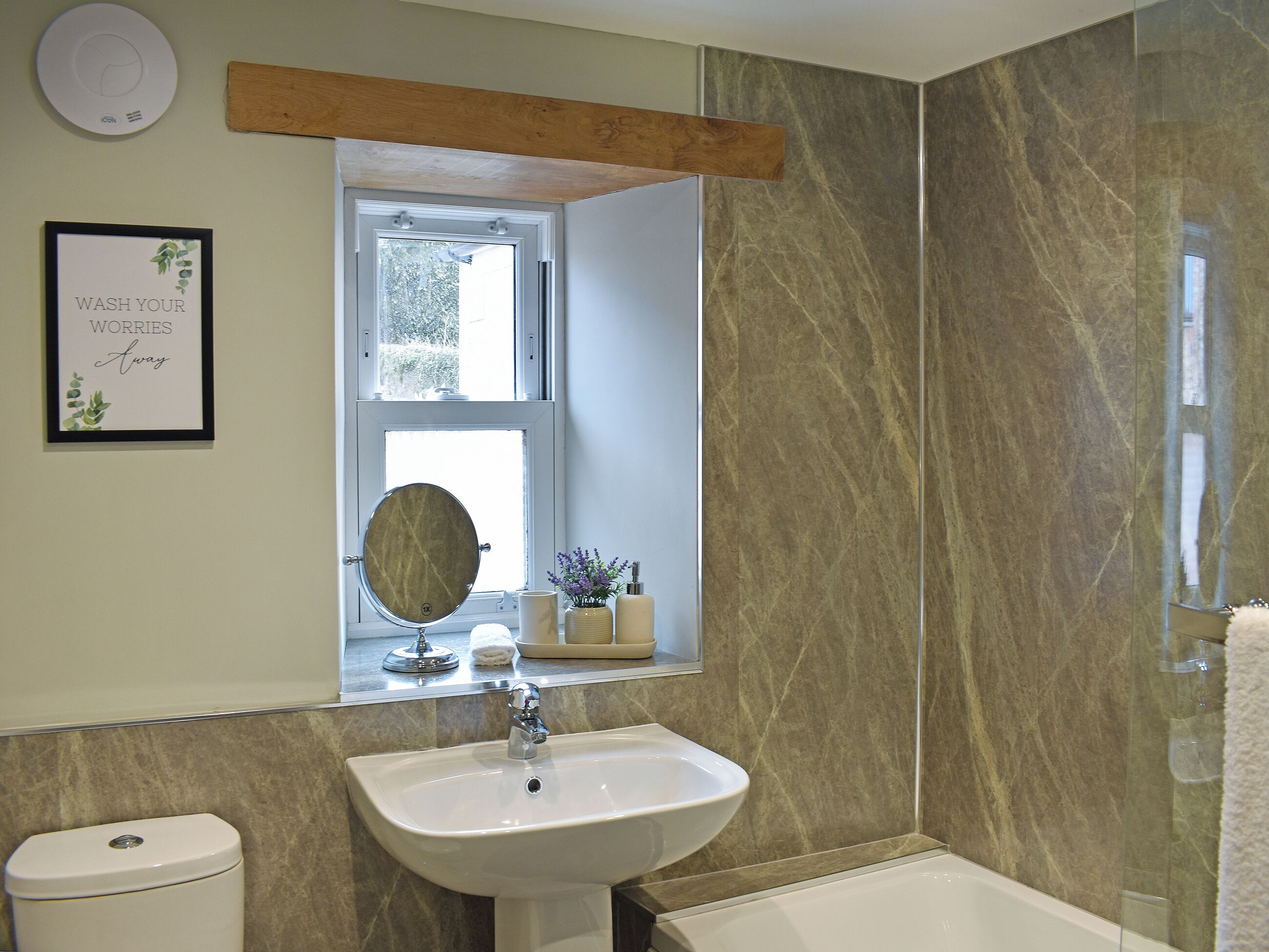 Ein Badezimmer mit Waschbecken, Toilette und Badewanne. Über dem Waschbecken befindet sich ein Fenster und ein Spiegel, an der Wand hängt ein gerahmtes Schild.