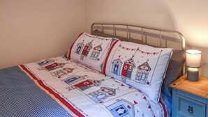 Un lit avec une couvre-lit à motifs et des oreillers, une table de chevet avec une lampe, et un mur orné d’un tableau.