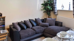 Un salon moderne avec un canapé gris foncé, une petite table d’appoint en bois, une plante en pot et un coin repas avec une table dressée pour deux personnes.