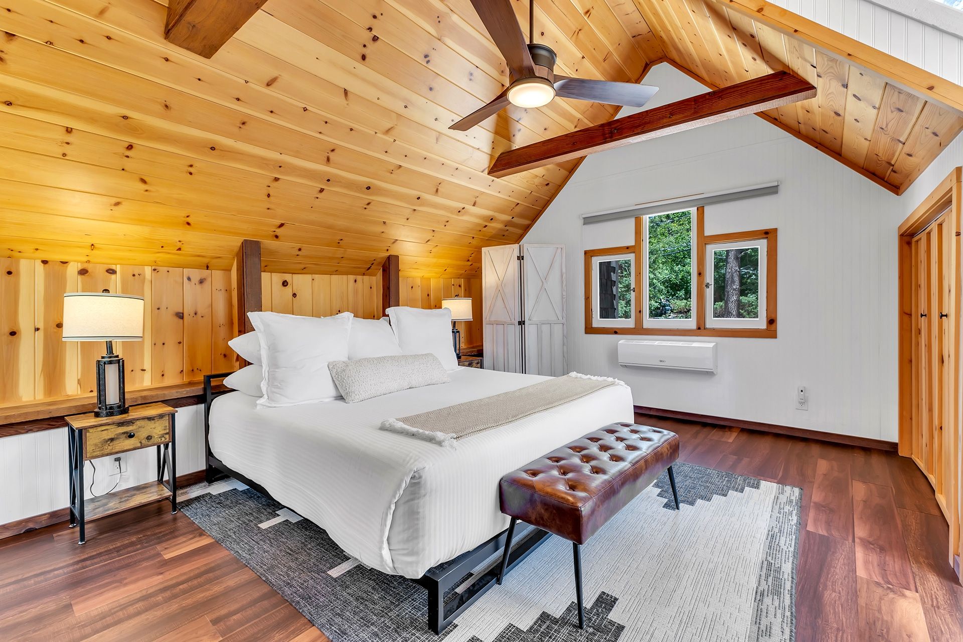 Une chambre avec un plafond en bois, un lit recouvert de draps blancs, un banc, une table de chevet et une fenêtre donnant sur des arbres.